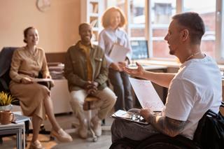 Man using a wheelchair in an office gestures to 3 onlookers, they are smiling.