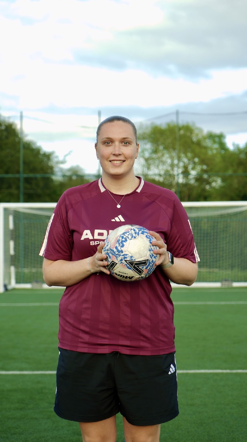 Lydia Shale standing on a football pitch smiling and holding a football.